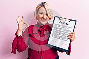 Young beautiful blonde plus size woman holding clipboard with contract document paper doing ok sign with fingers, smiling friendly