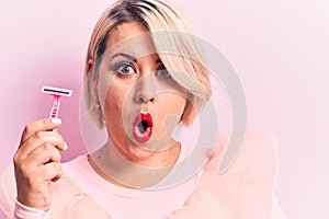 Young beautiful blonde plus size woman holding depilation razor over isolated pink background scared and amazed with open mouth