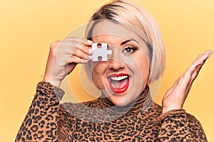 Young beautiful blonde plus size woman holding one piece of puzzle over eye celebrating achievement with happy smile and winner