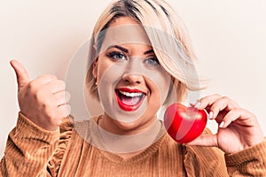 Young beautiful blonde plus size woman holding plastic heart over isolated white background pointing thumb up to the side smiling