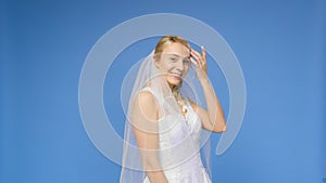 Young beautiful blonde smiling in a wedding white dress and veil on a blue background. The girl looks into the camera