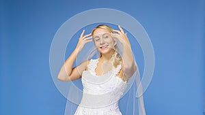 Young beautiful blonde smiling in a wedding white dress and veil on a blue background. The girl looks into the camera