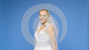 Young beautiful blonde smiling in a wedding white dress and veil on a blue background. The girl looks into the camera