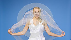 Young beautiful blonde smiling in a wedding white dress and veil on a blue background. The girl looks into the camera