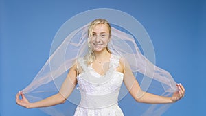 Young beautiful blonde smiling in a wedding white dress and veil on a blue background. The girl looks into the camera