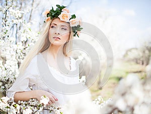 Young beautiful blonde smiling woman in white dress and rose wreath standing in blooming trees and looking aside