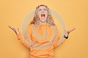 Young beautiful blonde sporty woman wearing casual sweatshirt over yellow background crazy and mad shouting and yelling with