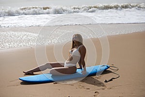 Young beautiful blonde surfer woman in white bikini sitting on blue surfboard. The girl enjoys her holidays on the beach to