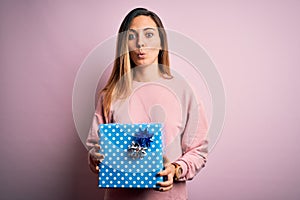 Young beautiful blonde woman with blue eyes holding birthday gift over pink background scared in shock with a surprise face,