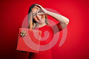 Young beautiful blonde woman with blue eyes holding birthday gift over red background with happy face smiling doing ok sign with