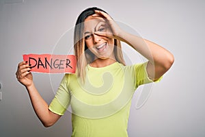 Young beautiful blonde woman with blue eyes holding paper with danger message with happy face smiling doing ok sign with hand on