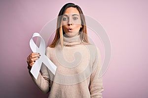 Young beautiful blonde woman with blue eyes holding white ribbon over pink background scared in shock with a surprise face, afraid