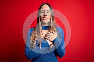 Young beautiful blonde woman with blue eyes standing over red background smiling with hands on chest with closed eyes and grateful