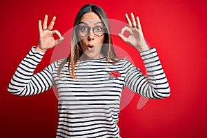 Young beautiful blonde woman with blue eyes wearing glasses standing over red background looking surprised and shocked doing ok
