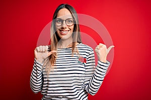 Young beautiful blonde woman with blue eyes wearing glasses standing over red background Pointing to the back behind with hand and