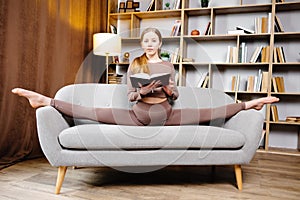 Young beautiful blonde woman in brown leggings and top sitting on a twine at home on the couch. The girl is reading a book sitting