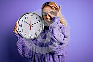 Young beautiful blonde woman doing countdown holding big clock over purple background with happy face smiling doing ok sign with