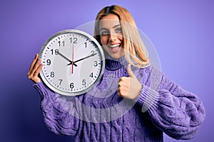 Young beautiful blonde woman doing countdown holding big clock over purple background happy with big smile doing ok sign, thumb up