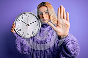 Young beautiful blonde woman doing countdown holding big clock over purple background with open hand doing stop sign with serious