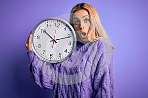 Young beautiful blonde woman doing countdown holding big clock over purple background scared in shock with a surprise face, afraid