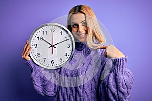 Young beautiful blonde woman doing countdown holding big clock over purple background with surprise face pointing finger to