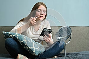 Young beautiful blonde woman doing makeup with make-up mirror and brush, sitting at home on bed, copy space