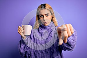 Young beautiful blonde woman drinking cup of coffee over isolated purple background pointing with finger to the camera and to you,