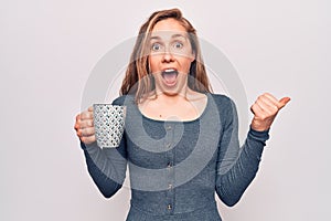 Young beautiful blonde woman drinking a cup of coffee pointing thumb up to the side smiling happy with open mouth