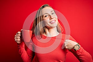 Young beautiful blonde woman drinking cup of coffee standing over isolated red background with surprise face pointing finger to