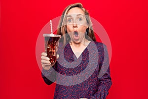 Young beautiful blonde woman drinking a fresh glass of soda scared and amazed with open mouth for surprise, disbelief face