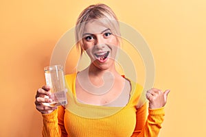 Young beautiful blonde woman drinking glass of water over isolated yellow background pointing thumb up to the side smiling happy