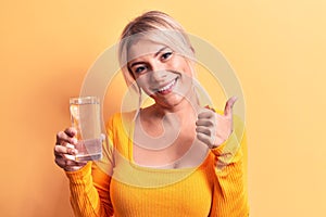 Young beautiful blonde woman drinking glass of water over isolated yellow background smiling happy and positive, thumb up doing