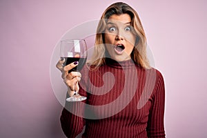 Young beautiful blonde woman drinking glasse of red wine over isolated pink background scared in shock with a surprise face,