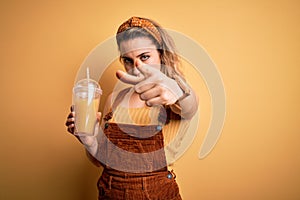 Young beautiful blonde woman drinking healthy glass of orange juice over yellow background pointing with finger to the camera and