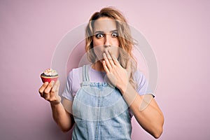 Young beautiful blonde woman eatimg chocolate cupcake over  pink background cover mouth with hand shocked with shame for