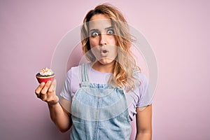 Young beautiful blonde woman eatimg chocolate cupcake over  pink background scared in shock with a surprise face, afraid