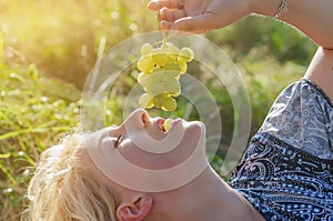 Young beautiful blonde woman eating bunch of grapes in nature in sun rays