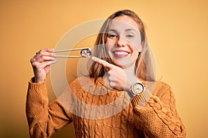 Young beautiful blonde woman eating sushi using chopsticks over isolated yellow background very happy pointing with hand and