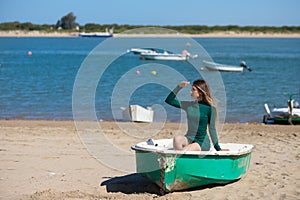 Young, beautiful blonde woman in an elegant green dress is sitting in a green fisherman's boat on the seashore. In the background