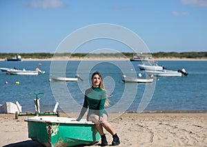 Young, beautiful blonde woman in an elegant green dress is sitting in a green fisherman's boat on the seashore. In the background