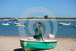 Young, beautiful blonde woman in an elegant green dress is sitting in a green fisherman\'s boat on the seashore. In the background