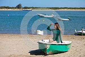 Young, beautiful blonde woman in an elegant green dress is sitting in a green fisherman\'s boat on the seashore. In the background