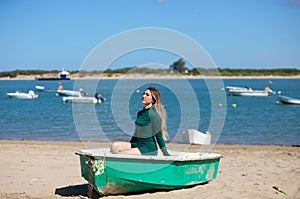 Young, beautiful blonde woman in an elegant green dress is sitting in a green fisherman\'s boat on the seashore. In the background