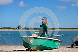 Young, beautiful blonde woman in an elegant green dress is sitting in a green fisherman's boat on the seashore. In the background