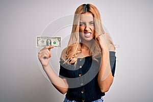 Young beautiful blonde woman holding 1 dollar banknotes over  white background annoyed and frustrated shouting with anger,