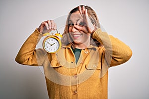 Young beautiful blonde woman holding alarm clock standing over isolated white background with happy face smiling doing ok sign