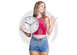 Young beautiful blonde woman holding big clock pointing thumb up to the side smiling happy with open mouth