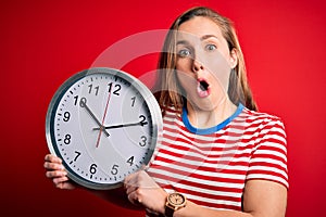 Young beautiful blonde woman holding big clock standing over isolated red background scared in shock with a surprise face, afraid