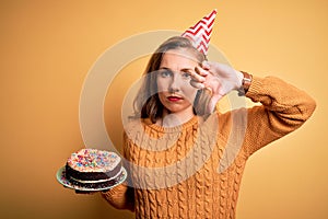 Young beautiful blonde woman holding birthday cake over isolated yellow background with angry face, negative sign showing dislike