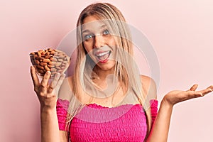 Young beautiful blonde woman holding bowl with almonds celebrating achievement with happy smile and winner expression with raised